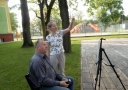 przed sztalugą na krześle stoi mężczyzna, obok niego stoi kobieta z uniesioną jedną ręką 