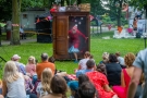 z przodu grupa ludzi siedząca na trawie, siedząca tyłem. Przed nimi aktor w czerwonym płaszczu i czapce, wychodzący z drewnianej szafy