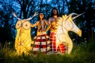 w nocnej scenerii, na trawie w tle drzewa, stoją dwaj przebrani aktorzy (kobieta i mężczyzna); po lewej stronie obok kobiety srtoi duży lampion przypominający sowę, po prawej stronie obok mężczyzny zaś lampion przypominający formą głowę smoka z grzywą i rogami
