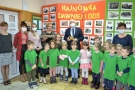 Dzieci, kadra pedagogiczna oraz delegacja z URzedu Miasta pozują do grupowego zdjecia. Z tyłu widać gazetkę ścienną ze zdjeciami Hajnówki dawniej i dziś