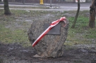Zdjęcie kamienia na którym jest przymocowana pamiątkowa tablica. Kamień owinięty jest biało - czerwoną wstążką.