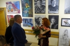 autorka wystawy stoi z kwiatami przed ekspozycją swoich prac. Dwie ososby gratulują jej udanej wystawy.