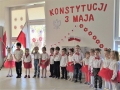 Zdjęcie przedstawia grupę dzieci w strojach biało czerwonych na tle dekoracji z okazji Konstytucji 3 Maja.