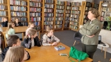 Dzieci siedzą przy stolikach  w czytelni i słuchają pracownika parku. 