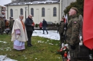 Proboszcz z organistą oddają hołd przy pomniku. składają przy pomniku wiązankę. Z prawej strony stoi żołnierz na warcie.