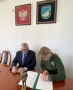 Prezes Fundacji podpisuje umowę, obok niej siedzi Burmistrz, na stole przed nimi stoi miniatura pomnika, 