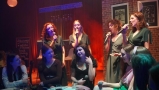 Dziewczęta siedzą przy stolikach.  Cztery z nich stoją z tyłu i śpiewają do mikrofonu. Sceneria jak w kawiarni.