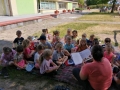 Dzieci siedzą i leżą na kocach w ogrodzie przedszkolnym, słuchają czytanych przez panie książek.