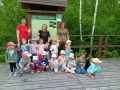 grupa dzieci wraz z opiekunami na tle tablicy edukacyjnej i zielonych drzew
