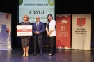 dwie kobieta i mężczyzna pozują do zdjecia na scenie. Kobieta po lewej trzyma czek z dotacją na 8 tys zł