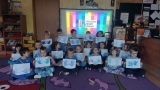 dzieci w niebieskich koszulkach trzymają w rękach kartki, na których są odciśniętymi na niebiesko dłońmi. W tle widać ekran z wyświetlanym slajdem na napiesem WSZYSCY MAMY PRAWA