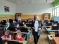dzieci siedza w ławkach szkolnych. przez śreodek przechodzi kobieta